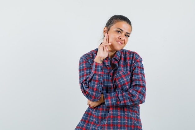 Mujer joven de pie en pose de pensamiento mientras sostiene la mano en la mejilla con camisa a cuadros y parece feliz. vista frontal.