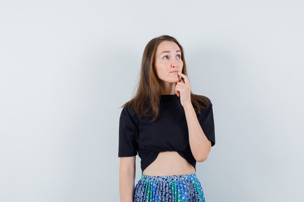 Mujer joven de pie en pose de pensamiento y mirando a otro lado en camiseta negra y falda azul y mirando pensativo