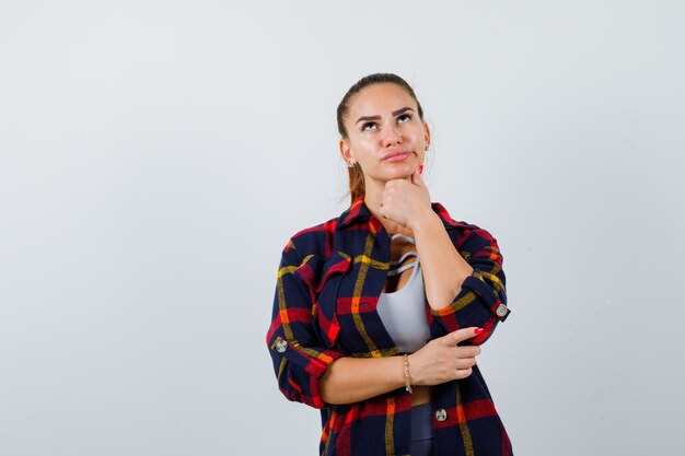 Mujer joven de pie en pose de pensamiento en la parte superior de la cosecha, camisa a cuadros y mirando pensativo, vista frontal.