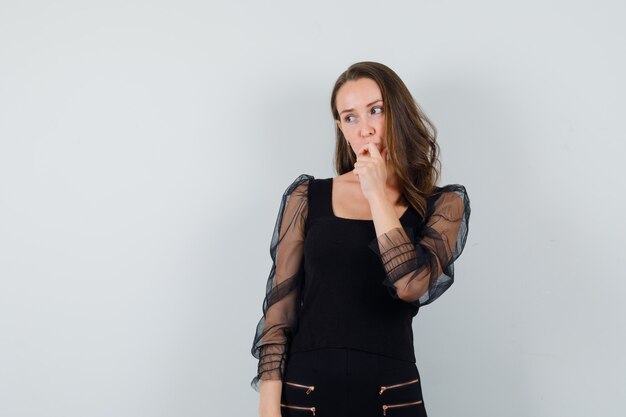 Mujer joven de pie en pose de pensamiento y poniendo el dedo índice en la boca en blusa negra y pantalón negro y mirando pensativo, vista frontal.