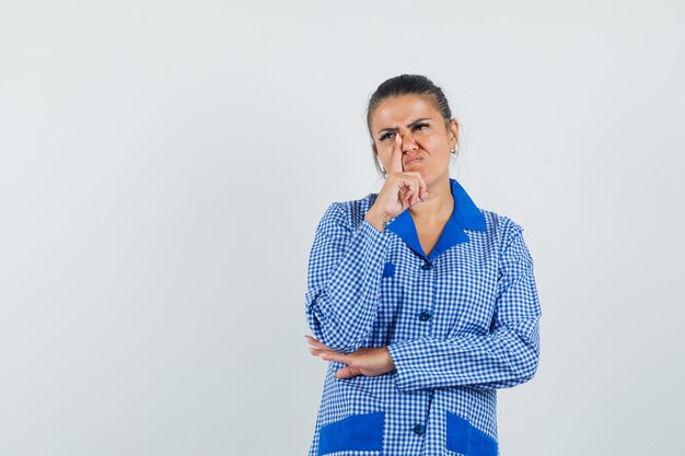 Mujer joven de pie en pose de pensamiento, poniendo el dedo índice cerca de la nariz en camisa de pijama de cuadros azules y mirando pensativo. vista frontal.