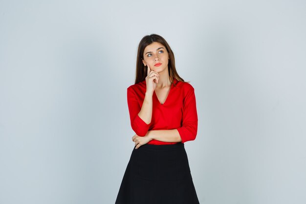 Mujer joven de pie en pose de pensamiento, poniendo el dedo índice debajo de la barbilla en blusa roja