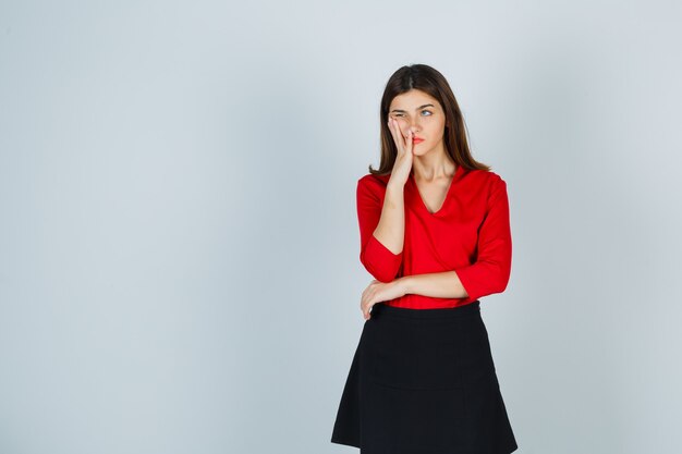 Mujer joven de pie en pose de pensamiento, poniendo la mano cerca de la boca en blusa roja