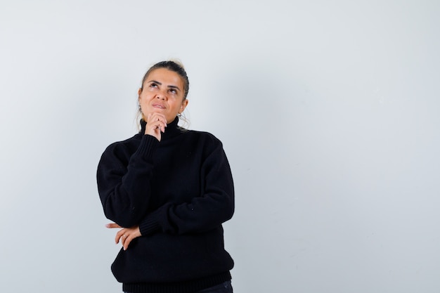 Mujer joven de pie en pose de pensamiento en suéter de cuello alto negro y mirando pensativo, vista frontal.