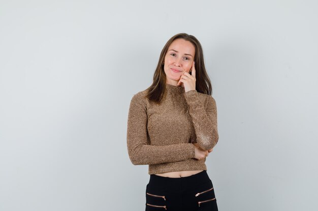 Mujer joven de pie en pose de pensamiento en suéter dorado dorado y pantalón negro y mirando feliz