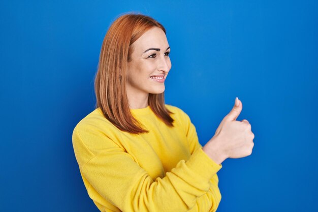 Mujer joven de pie sobre fondo azul luciendo orgullosa, sonriendo haciendo gestos de aprobación hacia un lado