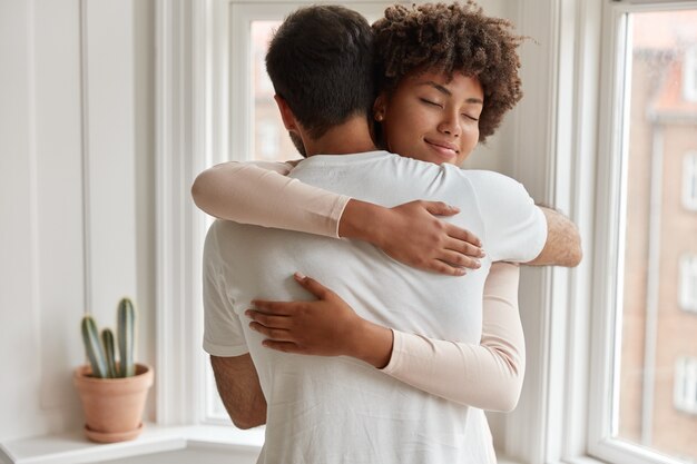 La mujer joven de piel oscura complacida le da un cálido abrazo a su novio, está complacida, posa junto a la ventana, tiene una relación romántica, se para en una habitación acogedora. Marido y mujer se sienten complacidos y unidos