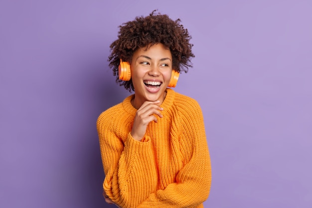 Mujer joven de piel oscura positiva escucha música en auriculares sostiene la barbilla y sonríe complacida concentrada a un lado se siente llena de alegría usa un cálido jersey naranja