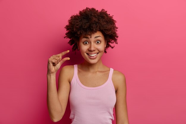 La mujer joven de piel oscura positiva satisfecha hace un letrero de tamaño pequeño, demuestra algo muy pequeño, hace gestos con la mano y da forma al objeto, usa chaleco informal, aislado sobre una pared rosa.