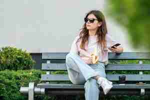 Foto gratuita una mujer joven con un plátano se sienta en un banco y usa un teléfono inteligente