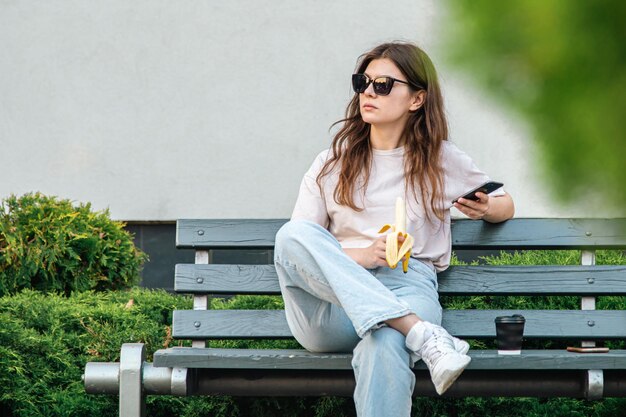 Una mujer joven con un plátano se sienta en un banco y usa un teléfono inteligente