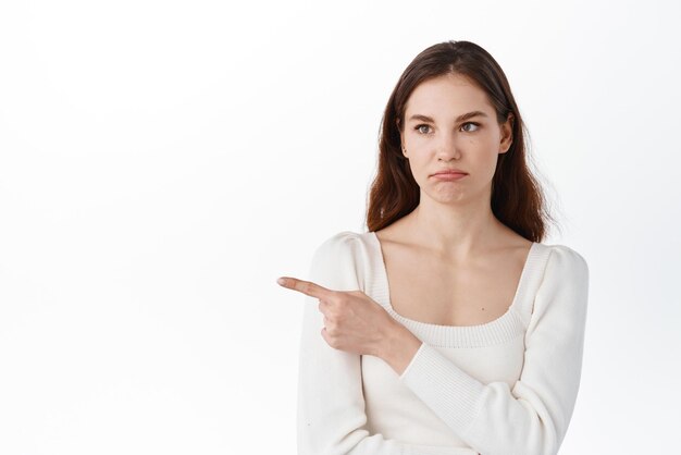 Mujer joven poco divertida y escéptica que señala con el dedo a un lado mirando a la izquierda con la cara disgustada y poco impresionada de pie contra el fondo blanco