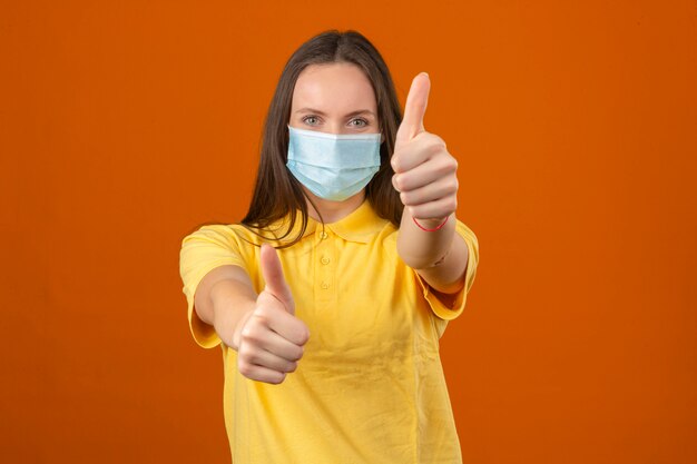 Mujer joven en polo amarillo y máscara de protección médica que muestra el pulgar hacia arriba signo mirando a la cámara con expresión positiva sobre fondo naranja