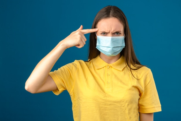 Mujer joven en polo amarillo y máscara protectora médica apuntando con el dedo a la cabeza con cara seria aislado sobre fondo azul.