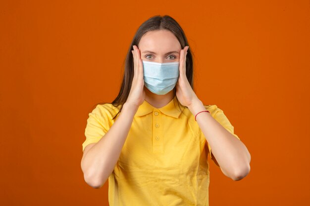Mujer joven en polo amarillo y máscara protectora médica tocando su rostro mirando a la cámara sobre fondo naranja