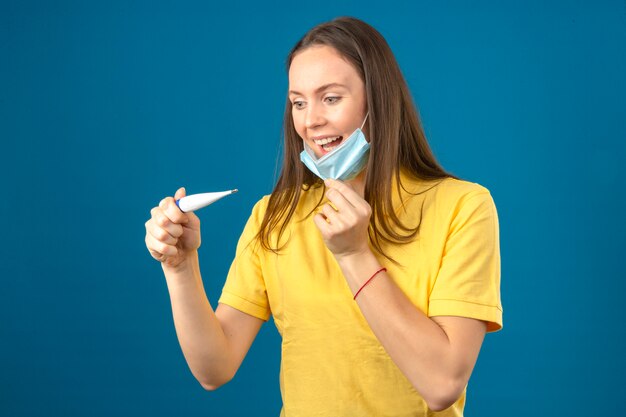 Mujer joven en polo amarillo quitándose la máscara protectora médica sonriendo mirando el termómetro con cara feliz aislado sobre fondo azul.