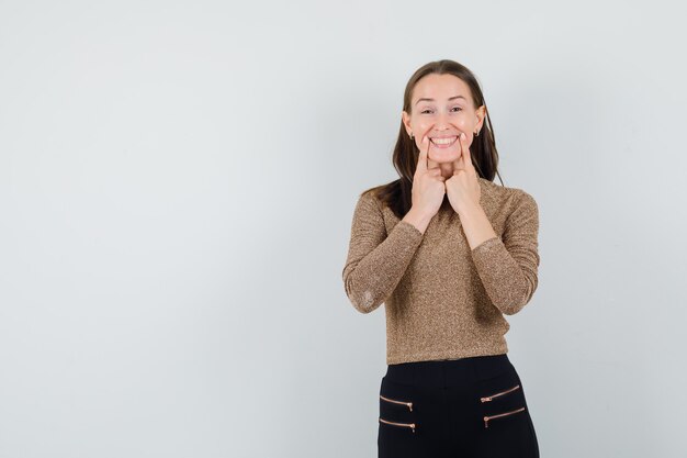 Mujer joven poniendo los dedos índices cerca de la boca, forzando una sonrisa en suéter dorado dorado y pantalón negro y luciendo feliz. vista frontal.