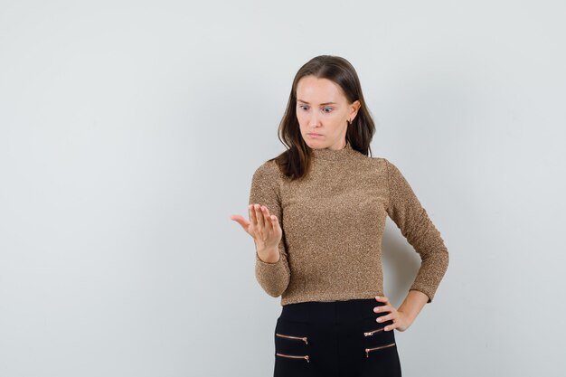 Mujer joven poniendo una mano en la cintura y mirando con la otra mano mientras sostiene el teléfono en suéter dorado dorado y pantalón negro y mirando sorprendido. vista frontal.