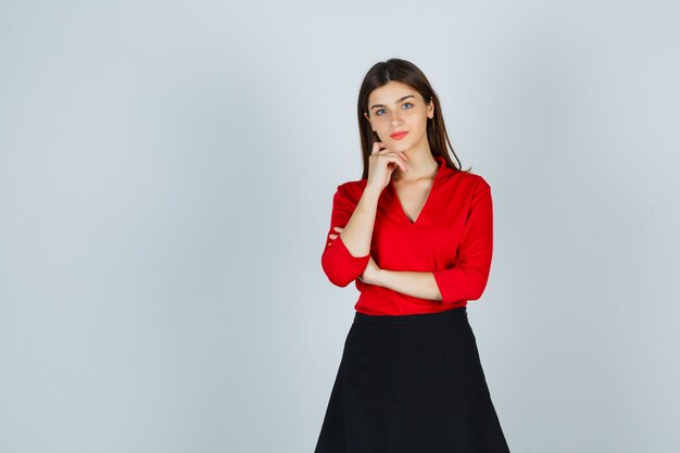 Mujer joven poniendo la mano debajo de la barbilla en blusa roja, falda negra y aspecto encantador