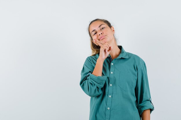 Mujer joven poniendo la mano debajo de la barbilla en blusa verde y mirando lindo