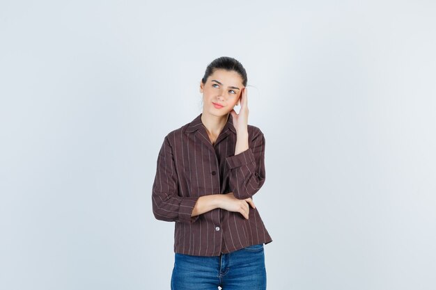 Mujer joven poniendo la mano en la sien, pensando en algo en camisa a rayas, jeans y mirando pensativo. vista frontal.
