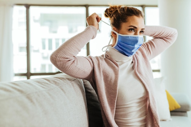 Mujer joven poniéndose una máscara facial mientras está en casa durante la epidemia de virus