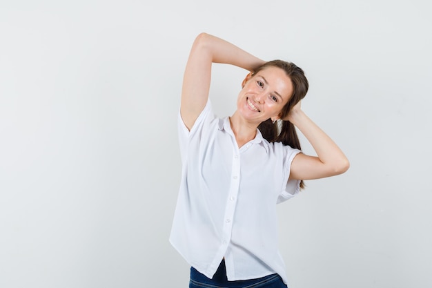 Mujer joven posando con la cabeza y los brazos en blusa blanca y mirando alegre.