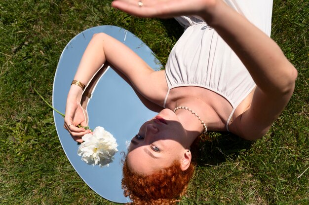 Mujer joven posando con flor de peonía en el espejo en el césped al aire libre