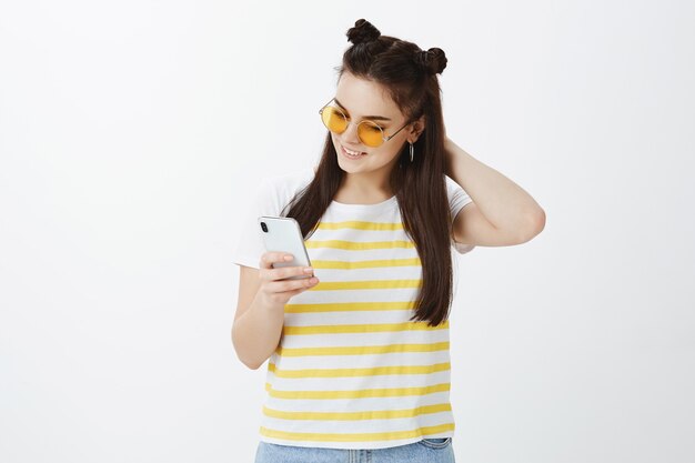 Mujer joven posando con gafas de sol y teléfono contra la pared blanca