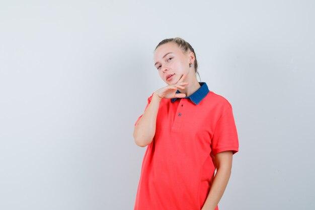 Mujer joven posando con la mano debajo de la barbilla en camiseta y luciendo elegante. vista frontal.