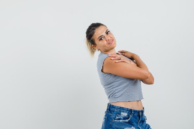 Mujer joven posando con las manos cruzadas sobre los hombros en camiseta, pantalones cortos y aspecto lindo.