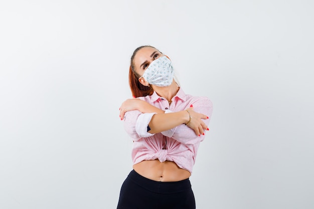 Mujer joven posando mientras se abraza a sí misma en camisa, pantalón, máscara y mirando soñadora, vista frontal.