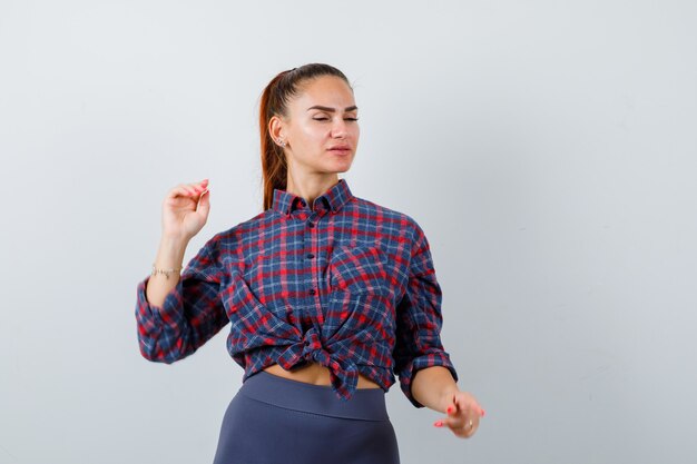 Mujer joven posando mientras está de pie en camisa a cuadros, pantalones y mirando confiado, vista frontal.