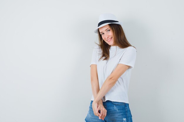 Mujer joven posando mientras está de pie en camiseta blanca, pantalones cortos, sombrero y mirando avergonzado.