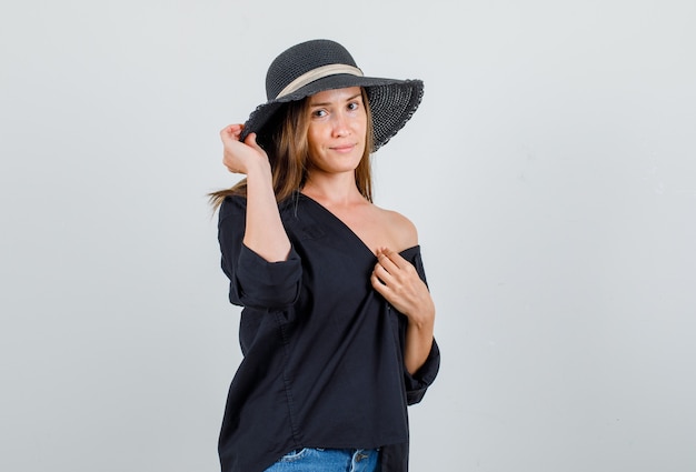 Mujer joven posando mientras mira a la cámara en camisa, pantalones cortos, vista frontal del sombrero.