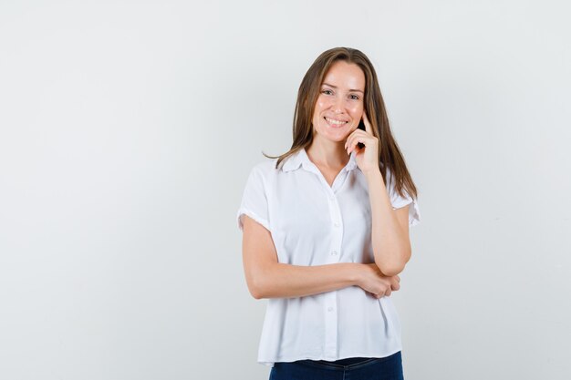 Mujer joven posando mientras sonríe en blusa blanca y parece positiva.