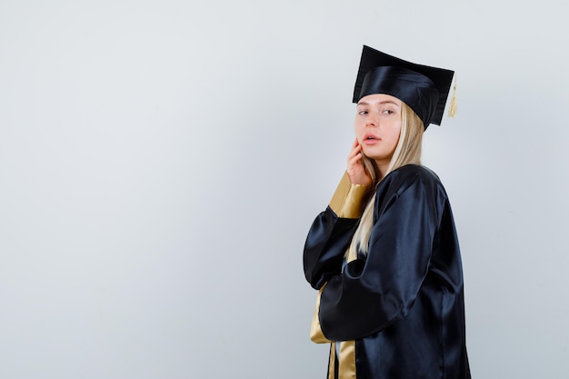 Mujer joven posando mientras toca su mejilla en uniforme de posgrado y luciendo seductora. .