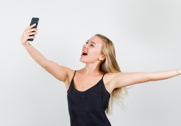 Mujer joven posando mientras toma selfie en camiseta negra y mirando alegre
