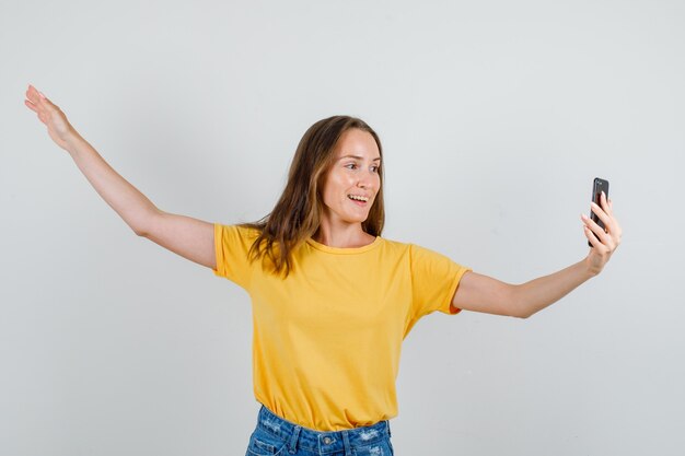 Mujer joven posando mientras toma selfie en camiseta, pantalones cortos y mirando alegre. vista frontal.