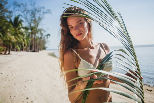 Mujer joven posando en la playa de arena bajo la hoja de palmera