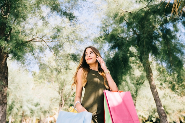 Mujer joven posando con teléfono y bolsas de compras en la naturaleza