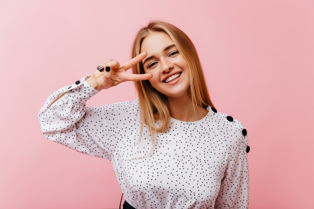 Mujer joven positiva en blusa de moda posando con el signo de la paz. Retrato interior de una encantadora mujer rubia sonriendo en rosa.