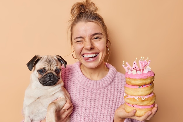 Mujer joven positiva y divertida guiña el ojo y saca la lengua tonteando mientras celebra el cumpleaños de las mascotas sostiene un perro pug y donas vestidas con un suéter de punto aislado sobre un fondo marrón