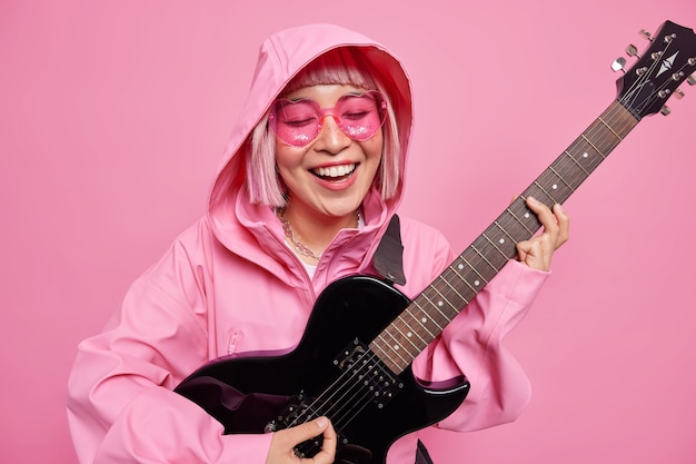 Mujer joven positiva entusiasta de la música toca música favorita sostiene la guitarra eléctrica sonríe felizmente estando de buen humor usa anorak con capucha en la cabeza gafas de sol de moda