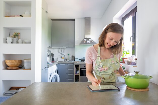 Mujer joven positiva escribiendo notas para la receta en la pantalla del pad, usando la tableta cerca de una cacerola grande en el mostrador. Vista frontal, espacio de copia. Cocinar en casa e internet concepto