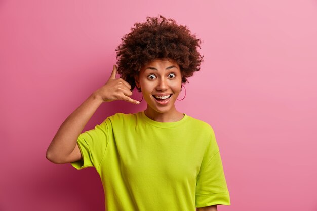 Mujer joven positiva hace un gesto de teléfono cerca de la oreja quiere tener una conversación contigo habla con una persona vestida con una camiseta informal posa contra la pared rosa