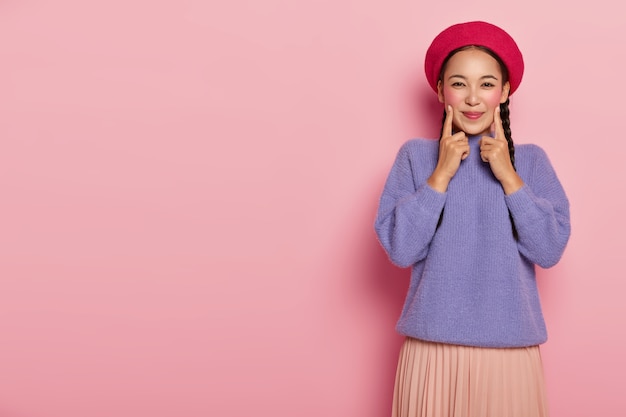 La mujer joven positiva mantiene los dedos índice en ambas mejillas, está muy animada, viste boina roja, suéter púrpura y falda, se encuentra sobre una pared rosa