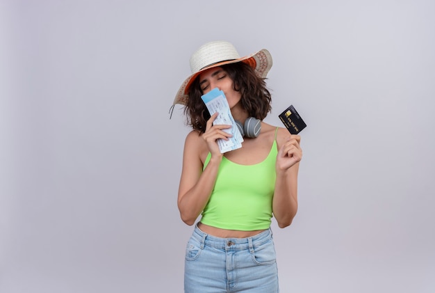 Una mujer joven positiva con el pelo corto en la parte superior de la cosecha verde en auriculares con sombrero para el sol besando boletos de avión con tarjeta de crédito sobre un fondo blanco.