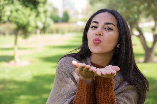 Mujer joven positiva que envía beso del aire en parque de la ciudad