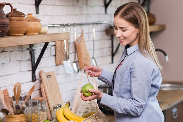 Mujer joven positiva que saca frutas orgánicas de la bolsa